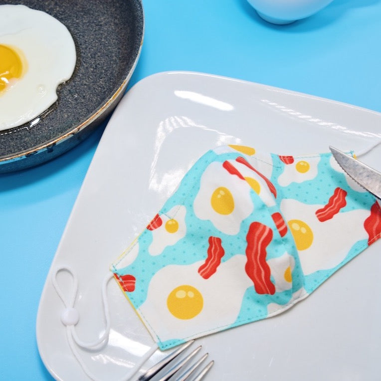 This adorable eggs and bacon mask features a combination of bold colors on a soft cotton print, forming a look that is both simple and fun. Made with one of our heavier-weight cotton novelty fabrics to provide a sturdier outer shell and the ultimate combination of both structure and comfort. Reversible and double-layered. Built with an adjustable nose bridge and adjustable ear straps.