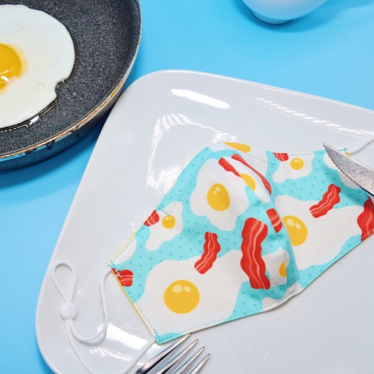 This adorable eggs and bacon mask features a combination of bold colors on a soft cotton print, forming a look that is both simple and fun.  Made with one of our heavier-weight cotton novelty fabrics to provide a sturdier outer shell and the ultimate combination of both structure and comfort. Reversible and double-layered. Built with an adjustable nose bridge and adjustable ear straps. 